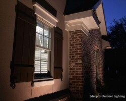 The image showcases a beautifully lit brick house exterior with strategic outdoor lighting, enhancing curb appeal and security during nighttime in Bluffton, SC. Outdoor window lighting in Bluffton illuminating the brick house exterior at night.
