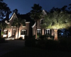 The image shows a stunning Beaufort home exterior brightly lit with outdoor lighting, highlighting the landscaping and architectural details at night. Beautifully illuminated Beaufort house exterior with landscape lighting and palm trees.