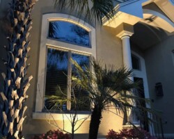 The image showcases a Hilton Head home with outdoor lighting design illuminating the facade, plants, and architectural elements in the evening. Outdoor house lighting in Hilton Head accentuates architectural details and landscape with warm, inviting illumination.